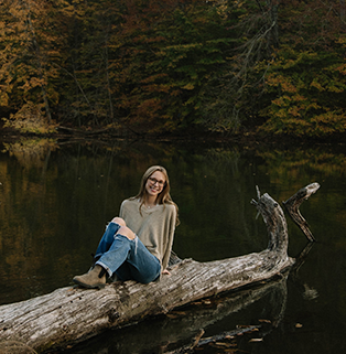 Photo of a white woman with glasses and light brown hair sitting on a log over water.